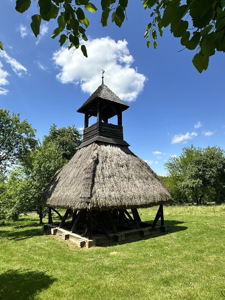 The thatched wooden belfry at Pankasz standing in a meadow under a blue sky
