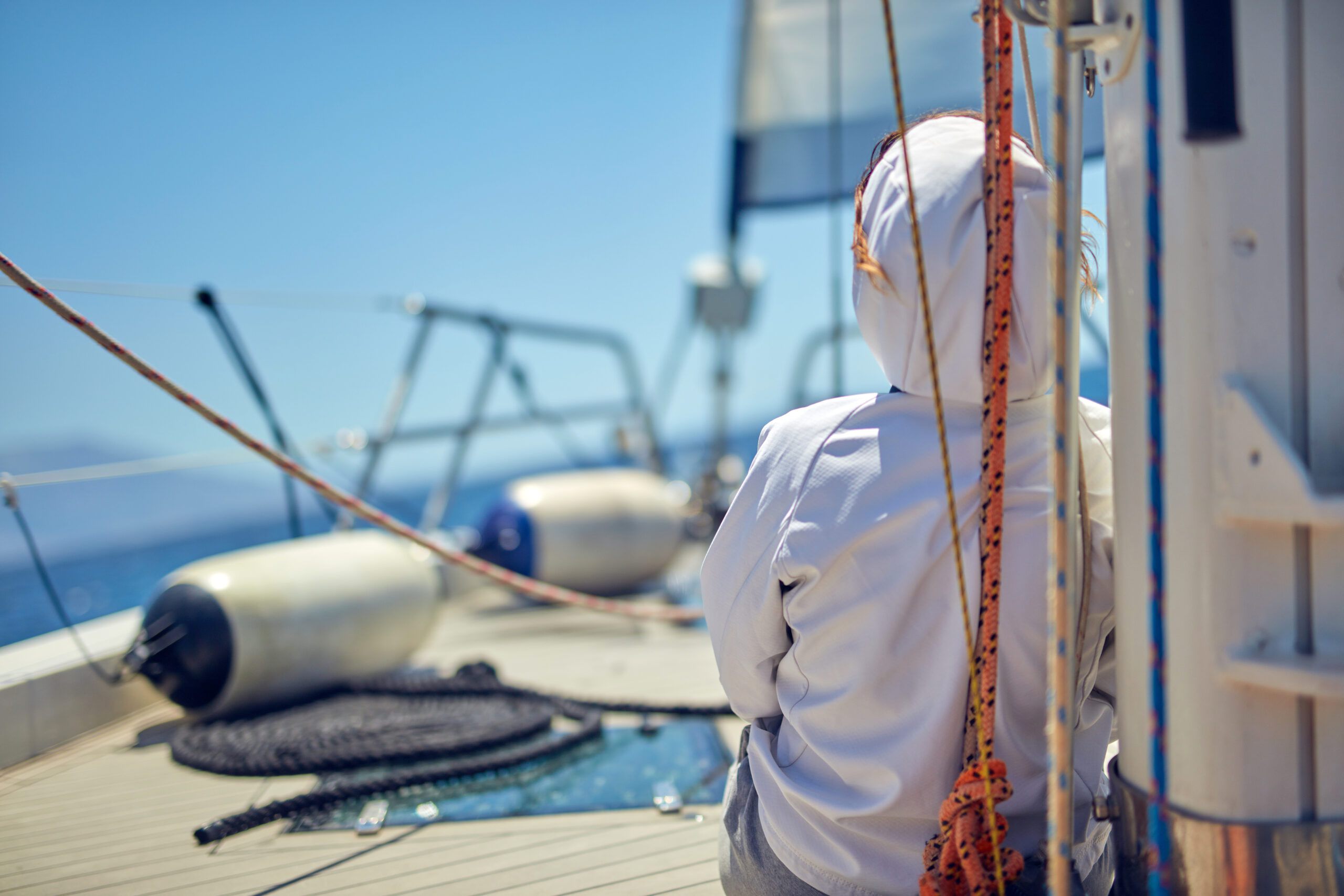 A sailor sitting on the foredeck of a yacht under sail, looking out over the water